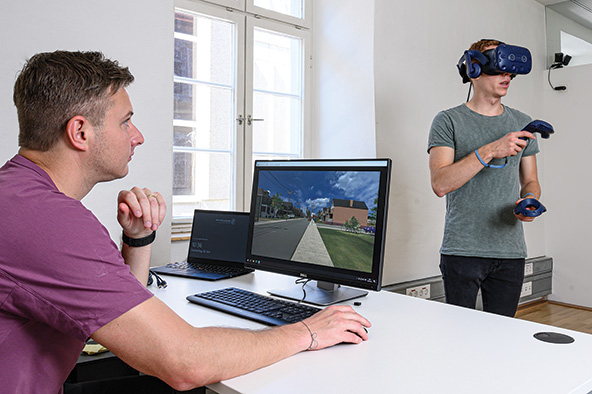 رجل يجلس أمام جهاز كمبيوتر ويراقب شخصاً يرتدي نظّارة الواقع الافتراضي