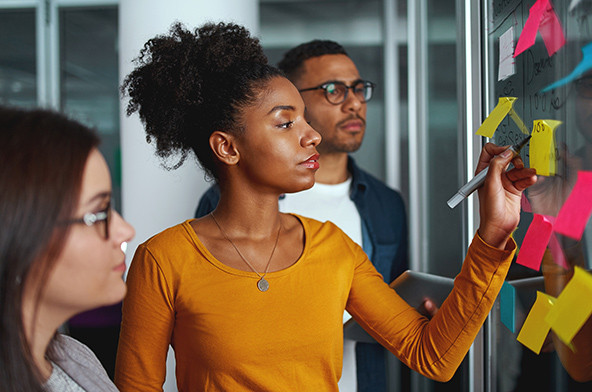 Young Black businesswoman standing with her colleagues writing new ideas on sticky notes over glass wall