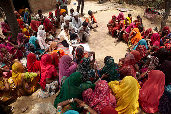 A group of people, many wearing colorful saris, sitting on the ground and listening to a presentation.
