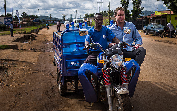 Two men riding a motorcycle that is pulling a cart with water jugs.