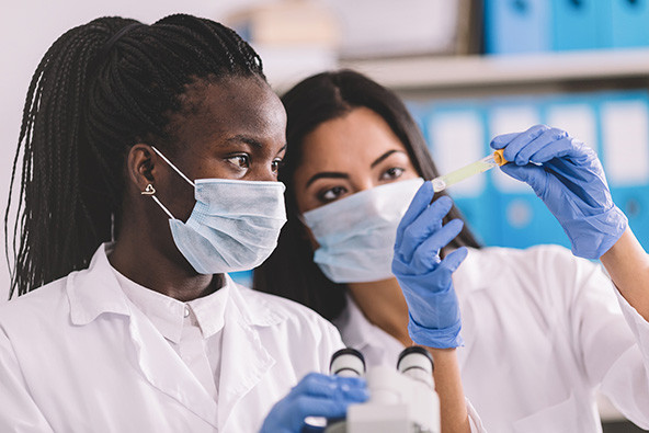 Two women wearing surgical masks, lab coats, and glove looking at a test tube.