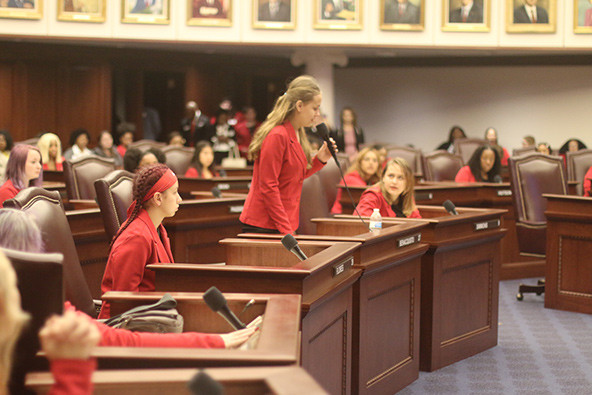 A young woman in a red jacket stands with a microphone at a desk in  a legislative chamber while other young women look on.