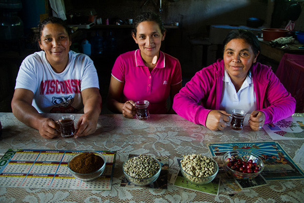 Three women coffee growers sitting at a wooden table with cups of coffee and four bowls of coffee beans in front for them.