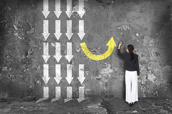 Difference thinking concept. A woman stands facing a wall. There are four columns of white arrows pointing down to her left. Her left arm is raised and shows her drawing a yellow arrow curving up, around, and toward the top of the wall.