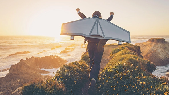 A young boy dressed in business suit and tie wears a homemade jetpack and flying goggles raises his arms in the afternoon sun while running to take off into the air.