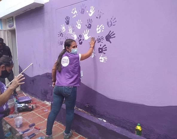 A woman wearing a purple T-shirt puts her painted hand on a wall to make her handprint.