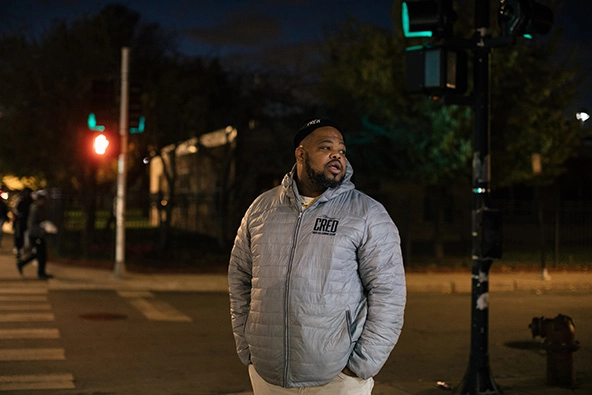 Man wearing a jacket with the letters CRED standing outside at night near a traffic light