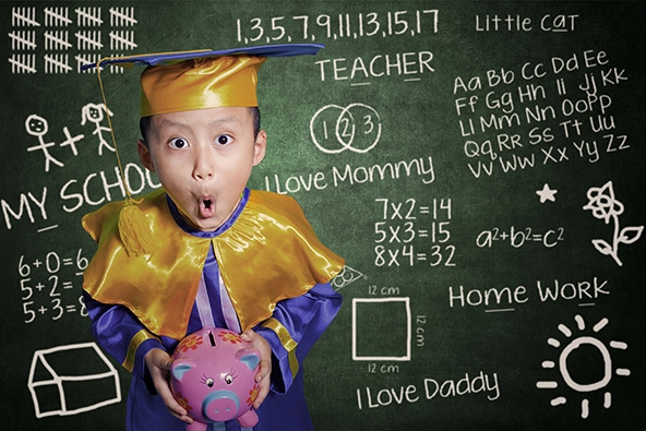 Child in graduation cap and gown holding a piggy bank and standing in front of a chalkboard