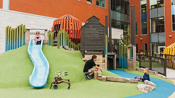 A child sits at the top of a slide and another child sits on the ground near a short wall made of wood on a therapeutic playground.