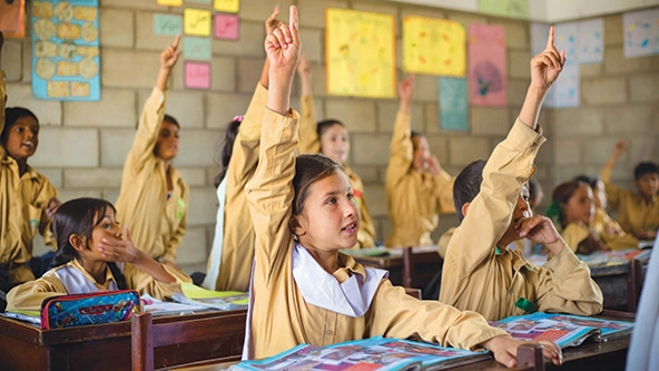 Young students in tan uniforms sitting in a classroom with hands raised.