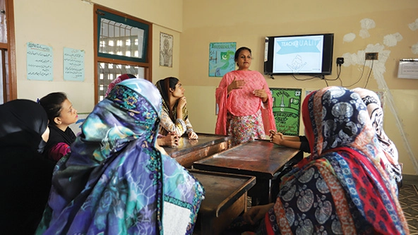 Woman stands at front of room with other teachers listening to her speak.