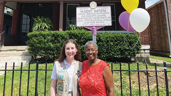 Young white woman and older African American woman stand in front of old house with a plaque that reads 