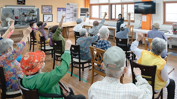 Elderly people sitting in chairs with one arm raised following instructions of person at front of room