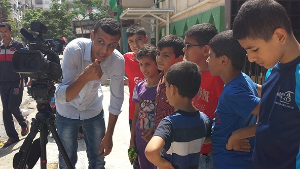 Palestinian man talking with young children points toward video camera