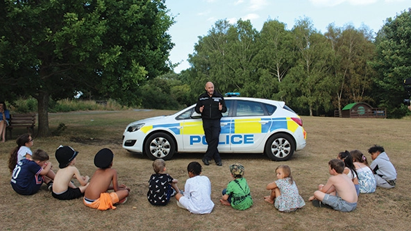 Police officer standing next to police car talks with semi-circle of seated children