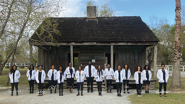 15 Black medical students in lab coats standing in front of a small wooden home on a plantation.