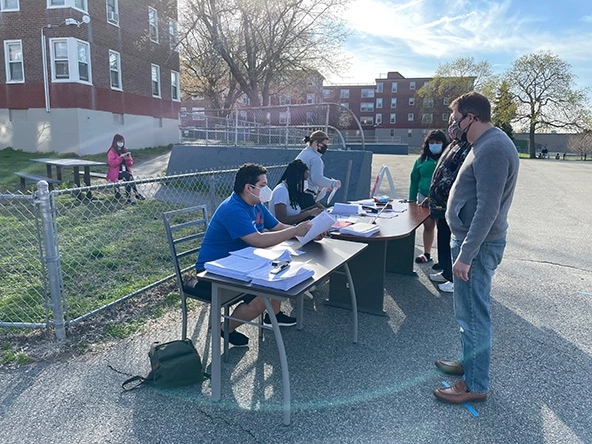 Three people lined up at two tables with three people behind tables handling paperwork. Everyone is outdoors and wearing surgical and non-surgical face masks.