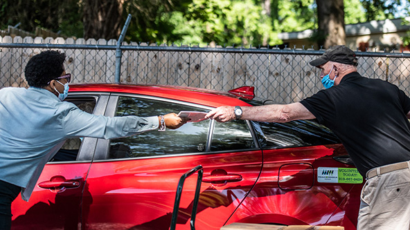 A Black woman wearing a face mask handing paperwork with an outstretched arm to a white man wearing a face mask with an outstretched arm. There is no contact between the two people.