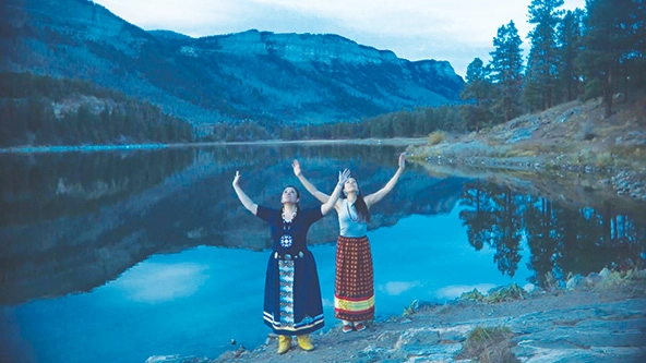 two native women standing on the shore of a lake with their arms stretched up to the sky