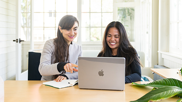 Two women sit at a table looking at a laptop together