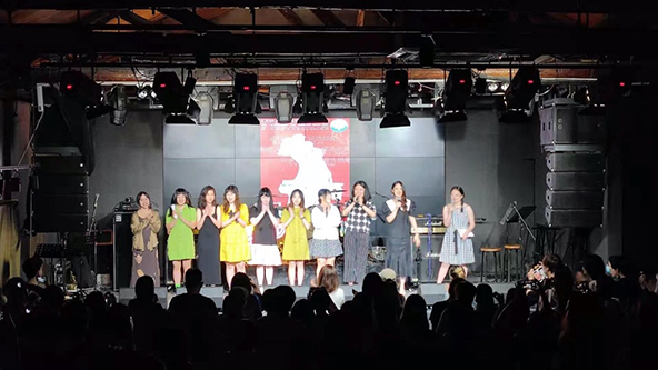 Women standing on a theater stage under lights in front of a crowd