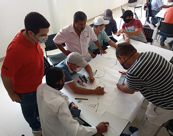 Eight men sitting and standing around a table are drawing a map of their community on a large piece of paper.