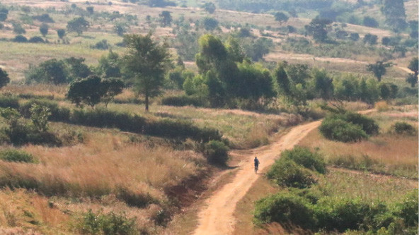 A young girl from Malawi riding a bicycle on a rural dirt road surrounded by grass, small shrubs, and trees.