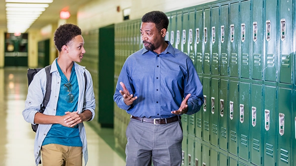 An African-American high school student walking with his teacher in the hallway by a row of lockers.