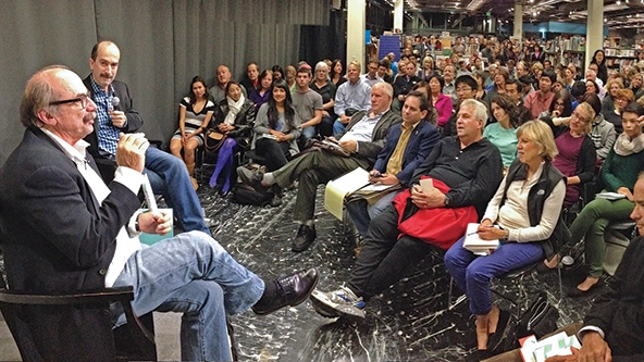 Two men on a stage talking to a large crowd of people inside a bookstore