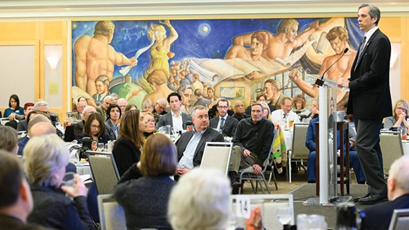 White man addressing a crowd seated at tables indoors