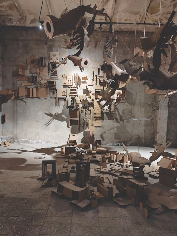 sculpture made of many tan to brown geometrical shapes and mannequin parts that is  suspended from the ceiling or resting on the floor in a museum