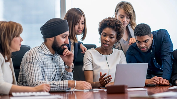A diverse group of business people gather around a laptop in a modern office