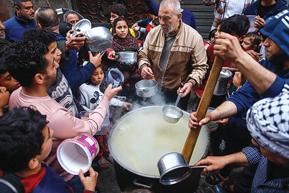 Palestinian man serving soup from a large pot surrounded by people