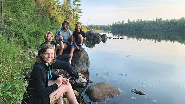 Youth sitting next to a river; one holding a fishing pole