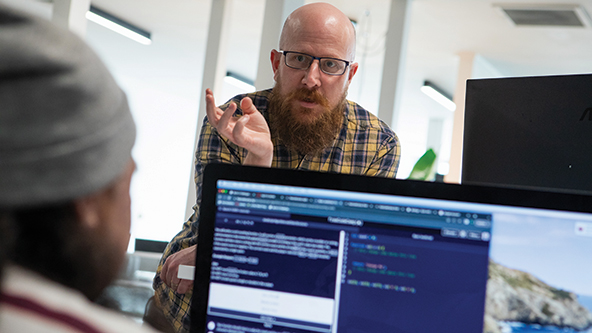 Bald man wearing glasses talks with a person working on a computer