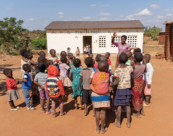 Woman raises her hand as a group of children watch her outside of a school in Malawi