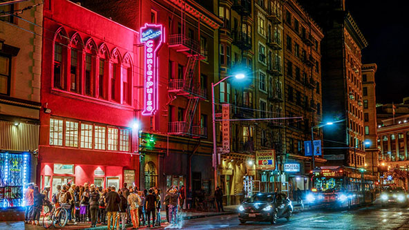 A crowd gathers on the street outside a building at night, illuminated by a neon pink sign that reads “San Francisco CounterPulse”
