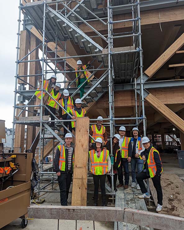 A group of men and women in neon yellow vests and construction hats smile at a construction site amid scaffolding.