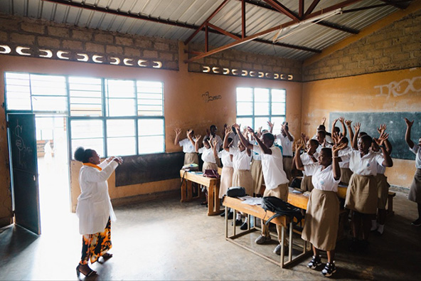 Rwandan teacher stands in front of classroom with students; everyone's arms are raised