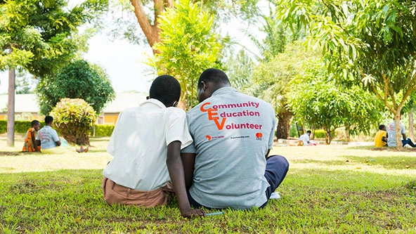 volunteer sits with student in the grass outside a school