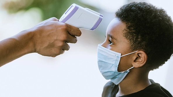 A hand holds an infrared thermometer up to a boy's forehead
